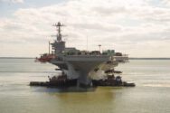 Navy nuclear aircraft carrier underway with tugs in a calm sea with cloudy skies