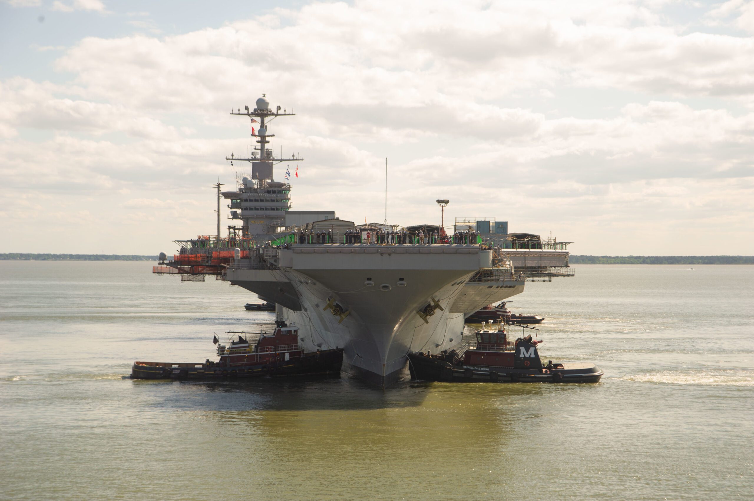 Navy nuclear aircraft carrier underway with tugs in a calm sea with cloudy skies