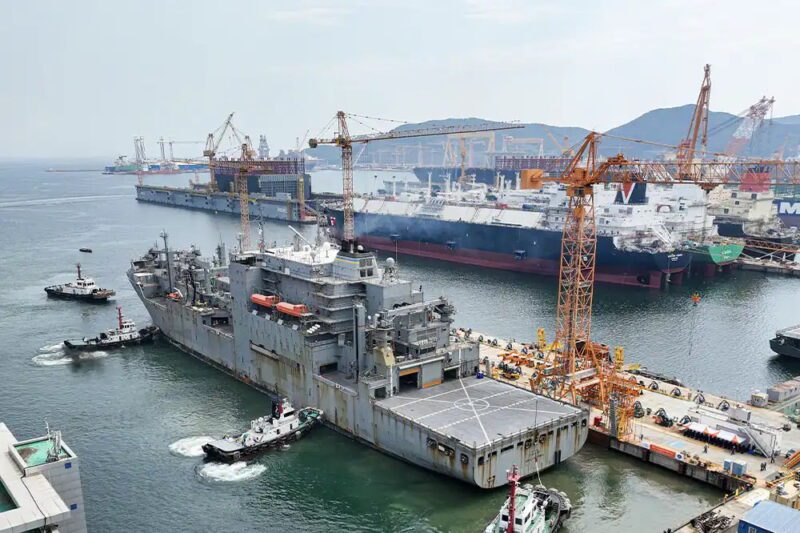 US Navy supply ship docking at a shipyard with tugboats