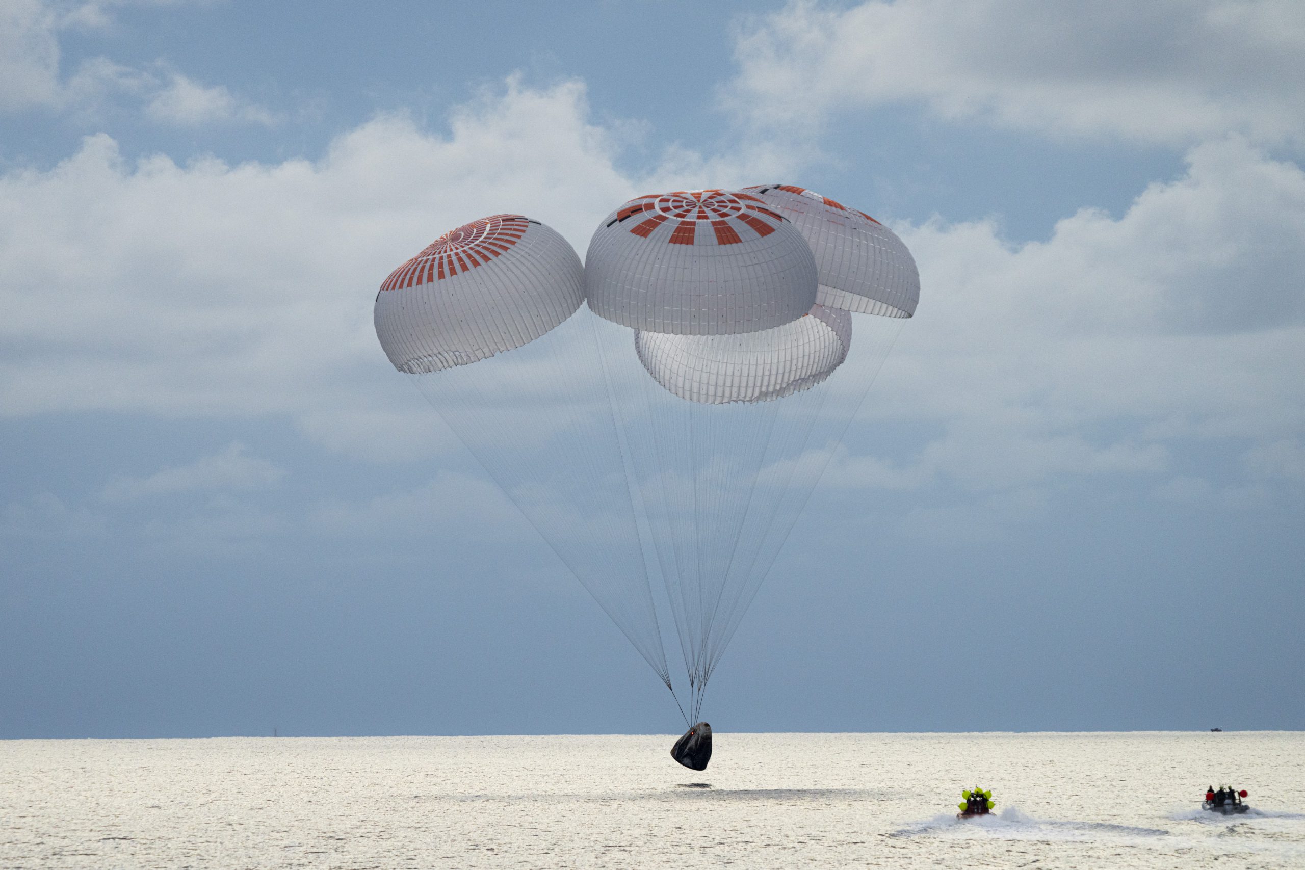 SpaceX Crew Dragon capsule descending with four large parachutes during atlantic ocean splashdown after completing the Polaris Dawn mission. Two recovery boats approach the capsule.