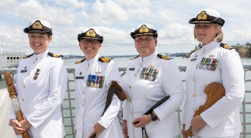 Image of fourfemale New Zealand Navy captains in full dress white uniforms holding M?ori tribal items with clouds and water in the background