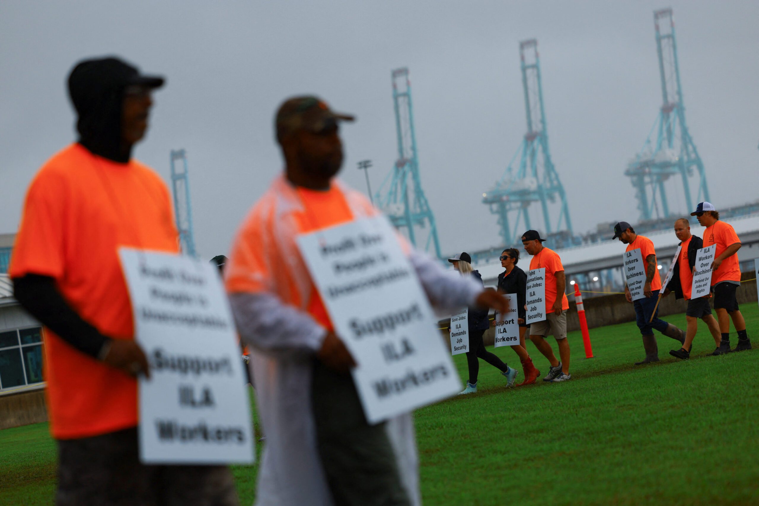 ILA picketers wearing orange safety shirts and carrying ILA signs with port cranes in the background