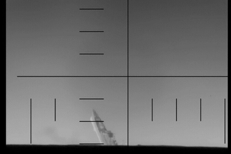 Thermal targeting view of an Iranian warship, identified as IRIS Dena, tilting steeply and sinking following a torpedo strike, seen through a crosshair targeting display at sea.
