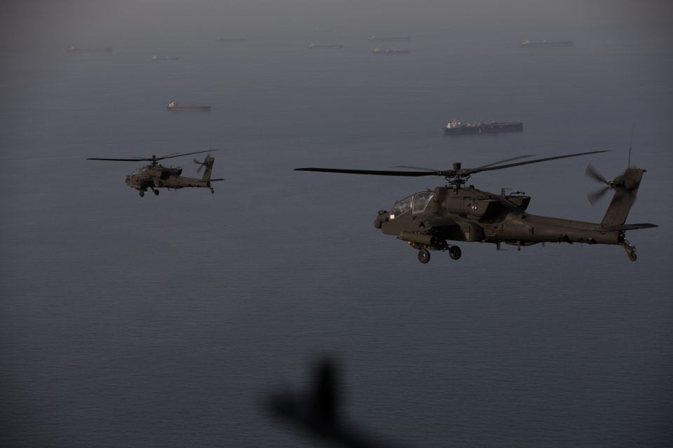 AH-64 Apache helicopters fly over the Strait of Hormuz, April 17, 2026, with multiple commercial vessels visible below, as U.S. Army crews maintain a persistent aerial presence to support freedom of navigation and monitor maritime traffic in the strategic waterway