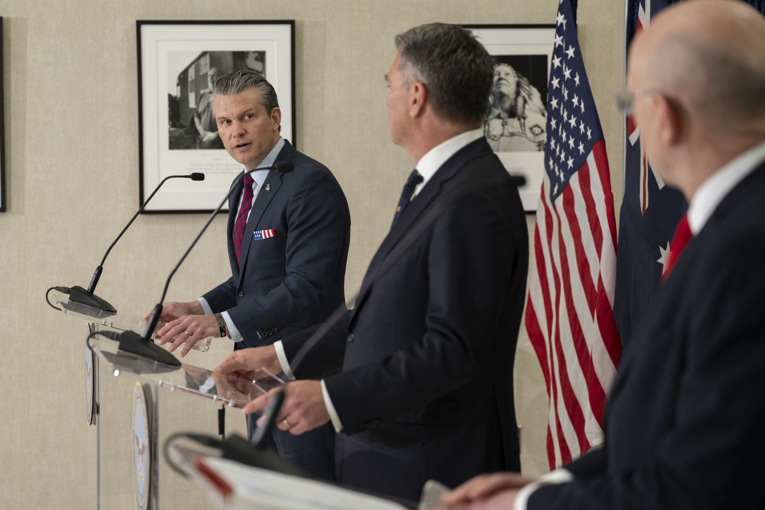 Senior U.S. and allied defense officials stand at podiums during a joint press event, with American and allied flags in the background. One speaker in a navy suit and patriotic pocket square turns toward another official mid-discussion, creating a candid moment of diplomatic exchange.