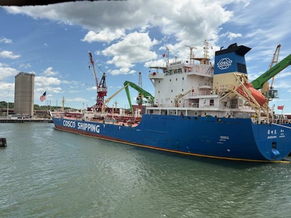 Chinese-flagged asphalt tanker JIN ZHOU WAN docked at a U.S. port. The blue COSCO Shipping vessel is moored alongside a terminal with cranes and industrial infrastructure visible in the background under partly cloudy skies.