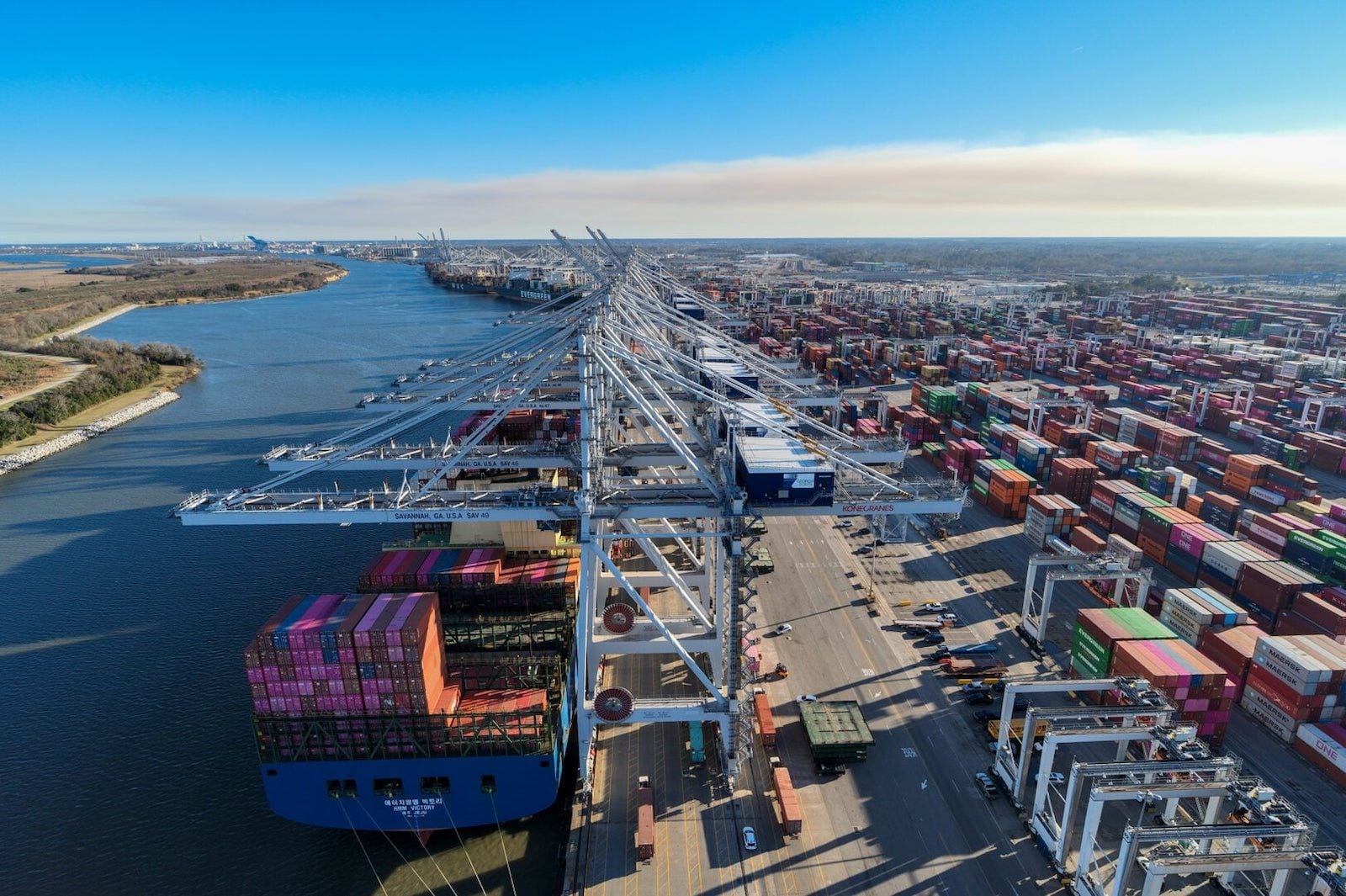 Aerial view of the Port of Savannah container terminal with massive ship-to-shore cranes, stacked shipping containers, and a container ship docked along the waterfront.