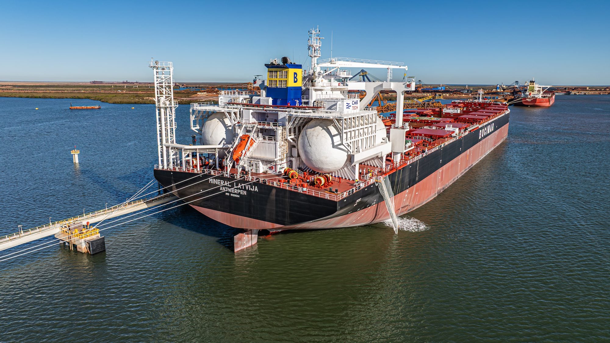 Aerial view of the ammonia-capable Newcastlemax bulk carrier Mineral Latvia maneuvering with tug assistance near an Australian export terminal. Large spherical ammonia fuel tanks are visible on the aft deck, highlighting the vessel's alternative-fuel design.
