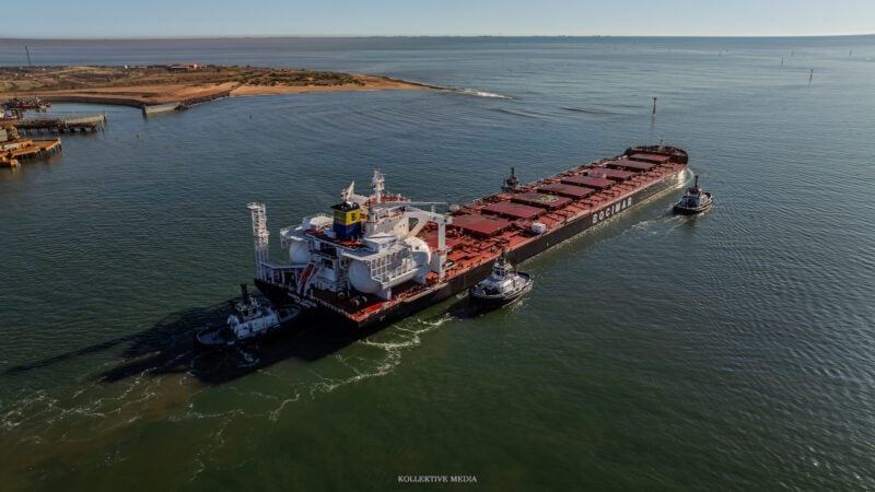 Aerial view of the ammonia-capable Newcastlemax bulk carrier Mineral Latvia maneuvering with tug assistance near an Australian export terminal. Large spherical ammonia fuel tanks are visible on the aft deck, highlighting the vessel's alternative-fuel design.
