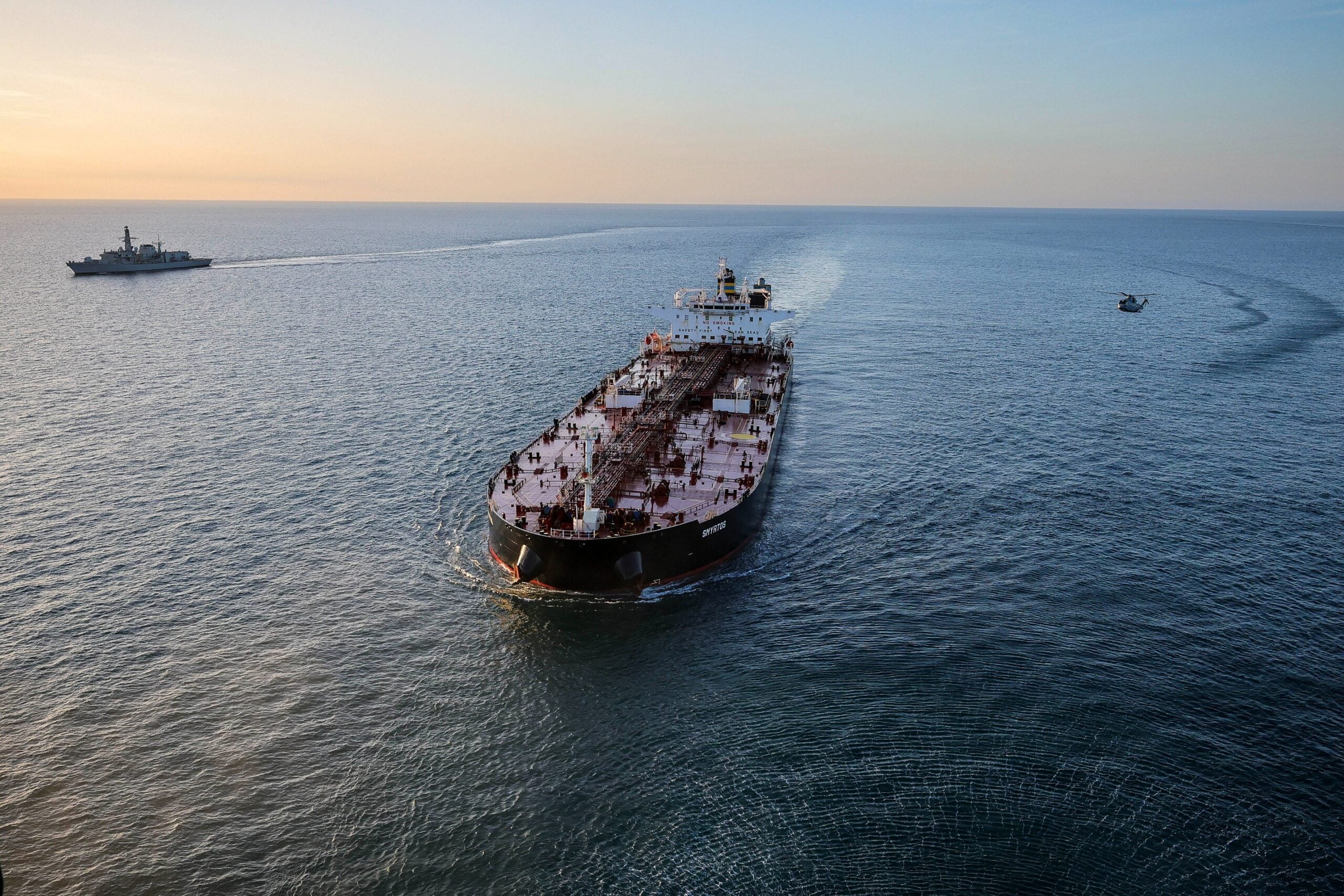 Aerial view of the tanker Smyrtos underway at sea, flanked at a distance by a Royal Navy warship and a helicopter following a UK enforcement operation in the English Channel.