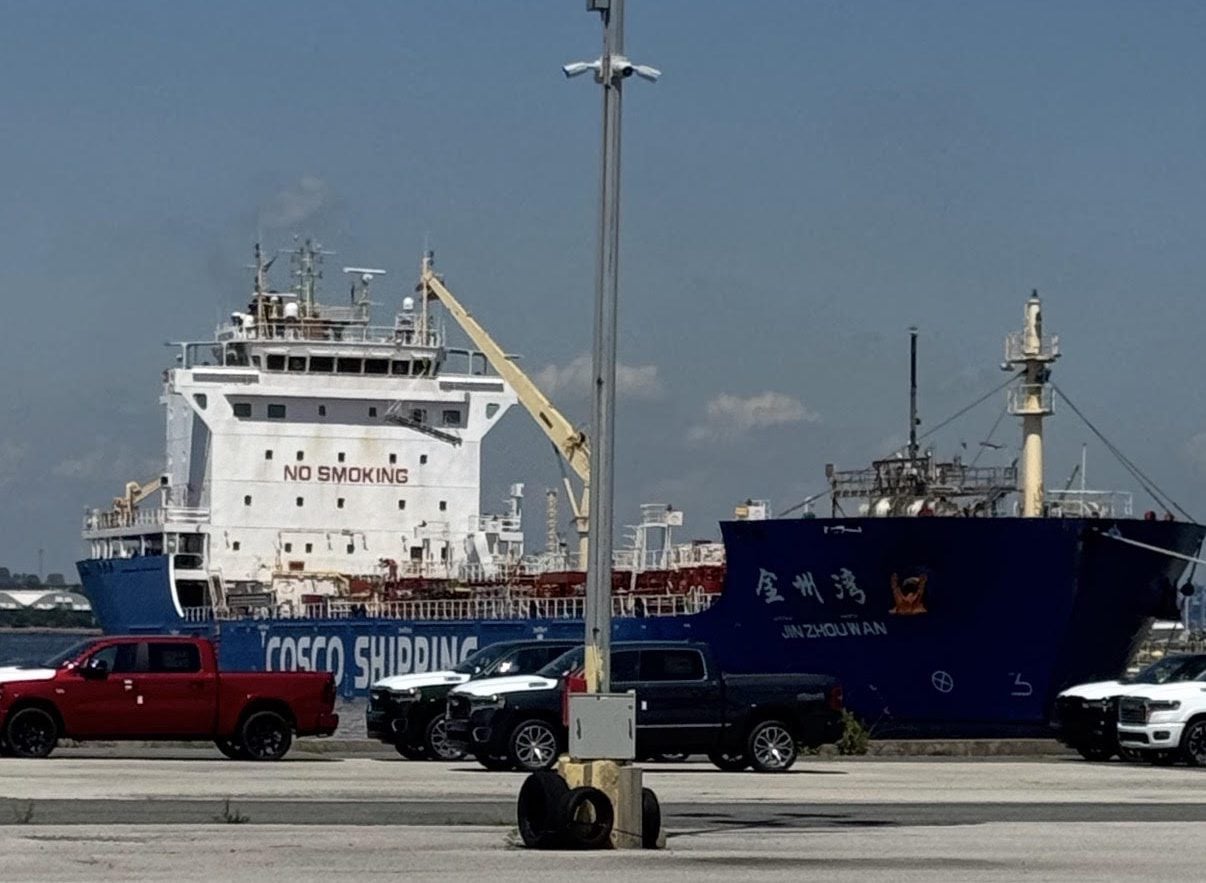 Chinese-flagged asphalt tanker JIN ZHOU WAN berthed in Baltimore after transporting asphalt from New Jersey under the emergency Jones Act waiver. The blue COSCO Shipping vessel is docked behind parked vehicles along the waterfront.