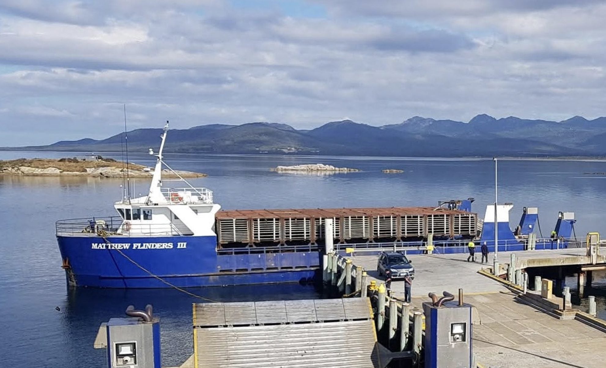 The cargo vessel Matthew Flinders III moored at a Tasmanian wharf with mountains and calm water in the background.