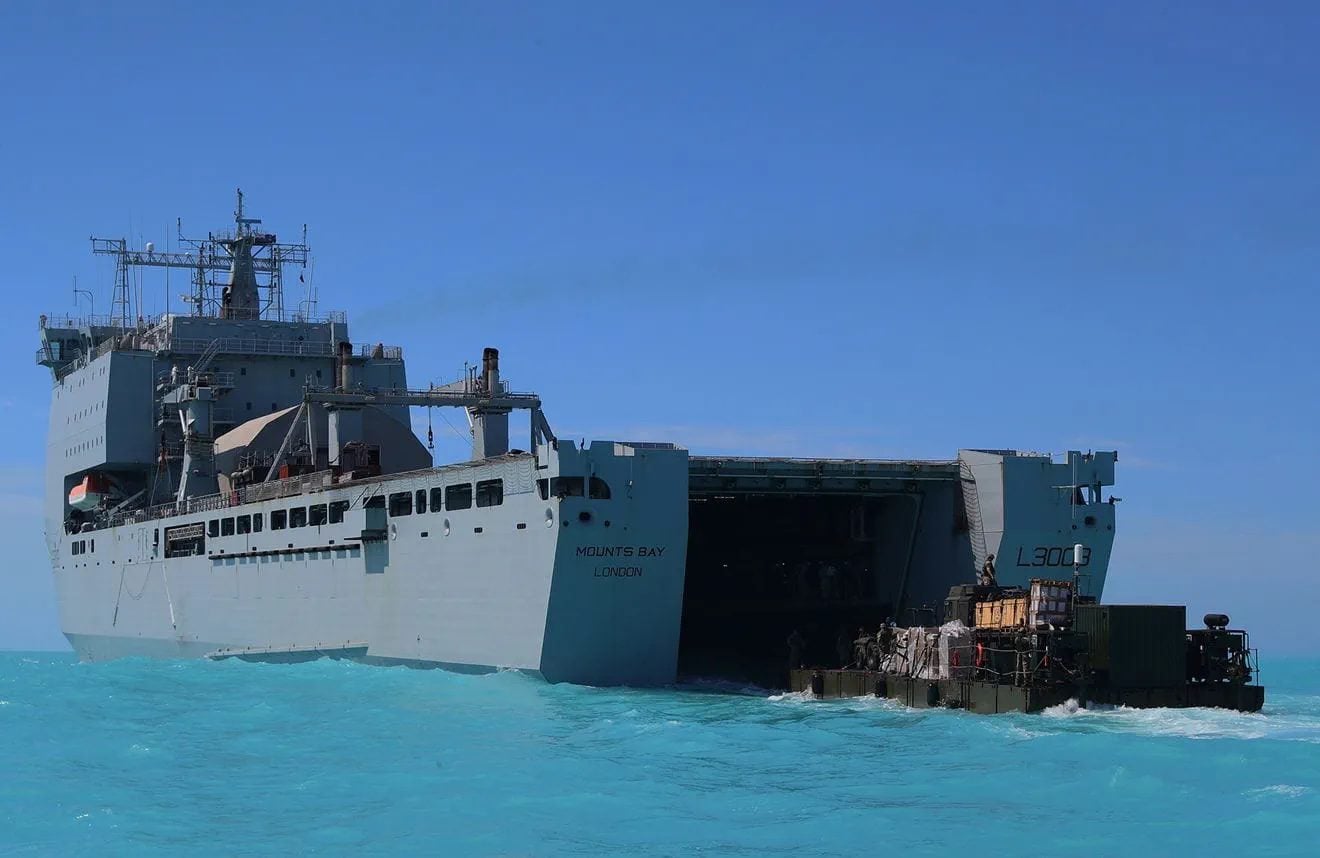 RFA Mounts Bay with its stern dock open as military vehicles and cargo are unloaded onto a landing craft in shallow turquoise water during an amphibious logistics operation.