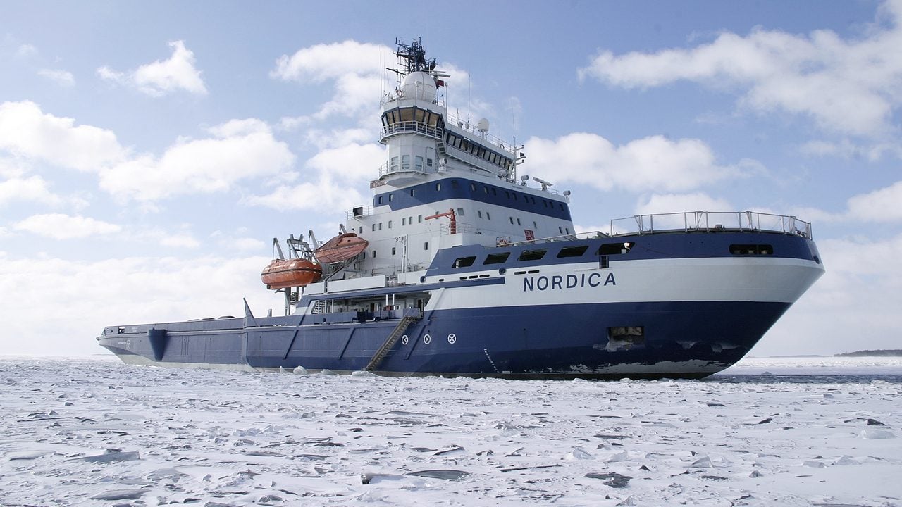 The Finnish multipurpose icebreaker Nordica navigates through sea ice, viewed from the starboard bow under a partly cloudy sky.