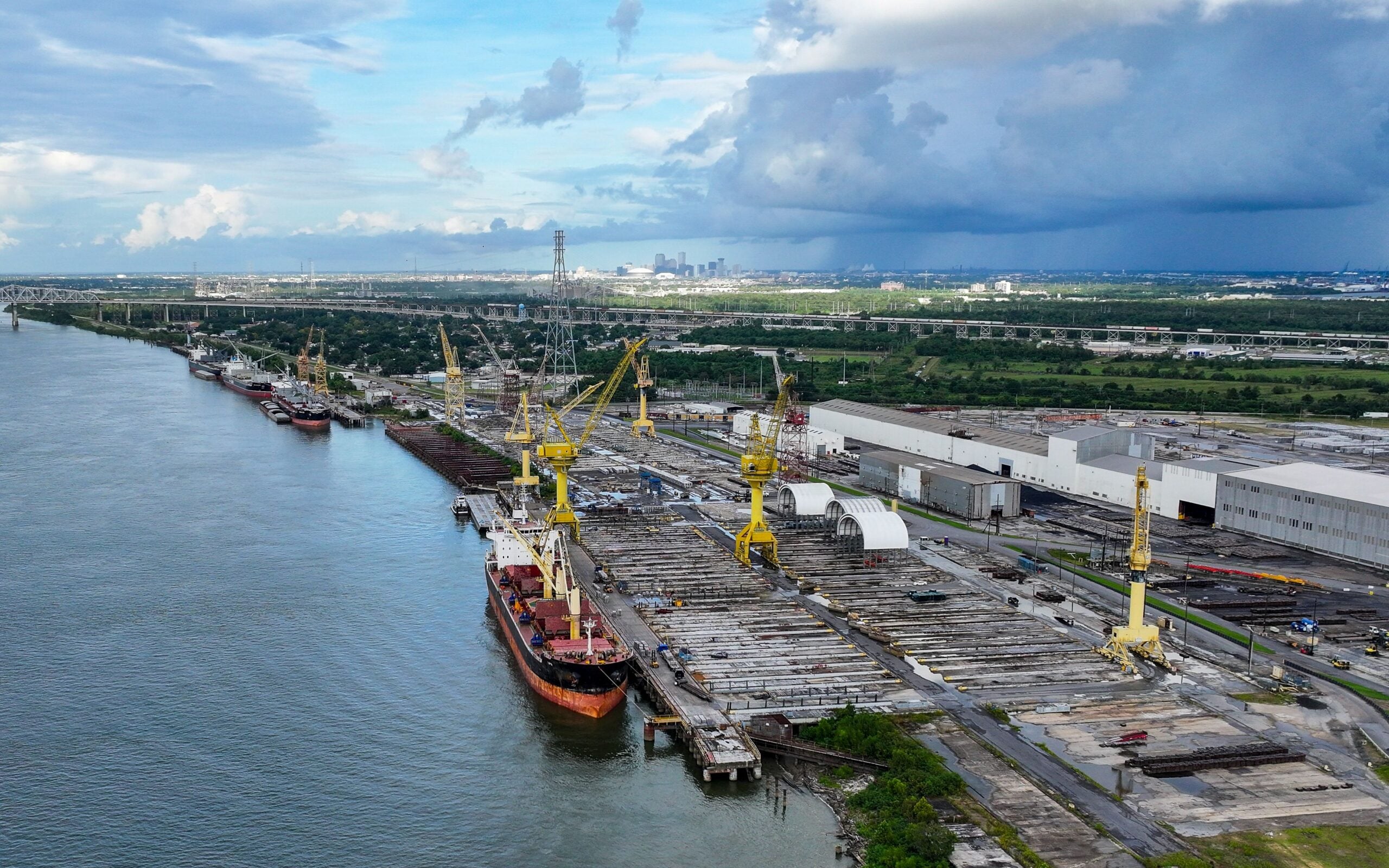 Aerial view of Avondale Global Gateway on the Mississippi River, showing cargo ships docked alongside large cranes, industrial warehouses and cargo storage areas, with New Orleans visible in the distance.