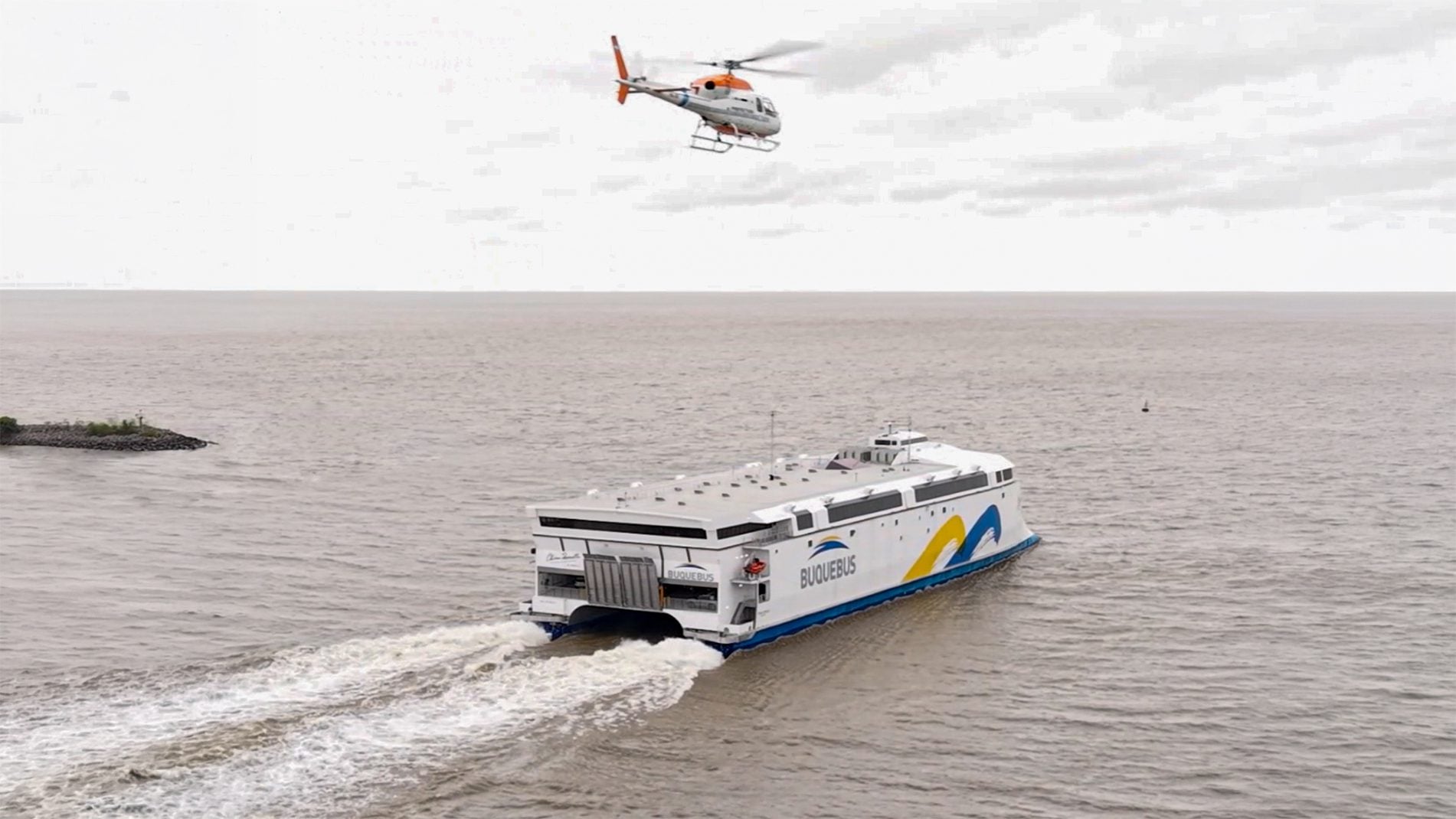 Aerial view of the 130-meter battery-electric ferry China Zorrilla underway on the River Plate, leaving a wake behind it as a helicopter flies overhead.